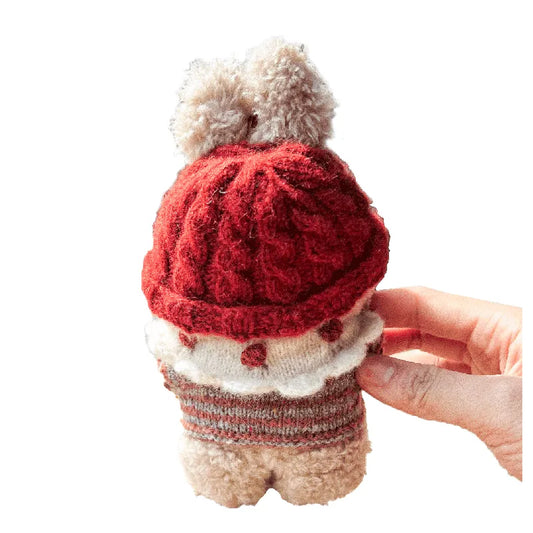 Small plush toy wearing a red knitted hat and sweater, held by a hand on a white background.