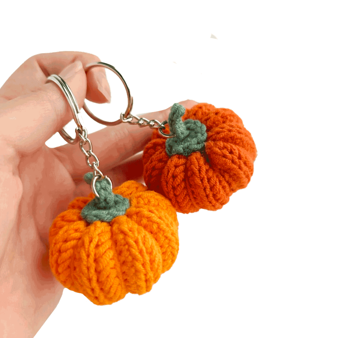 Hand holding two orange knitted pumpkin keychains with green stems on a white background