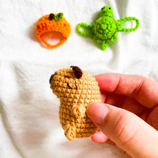 Hand holding a small crocheted capybara with green turtle and orange pumpkin crochet toys in background crochet mini capybara pattern