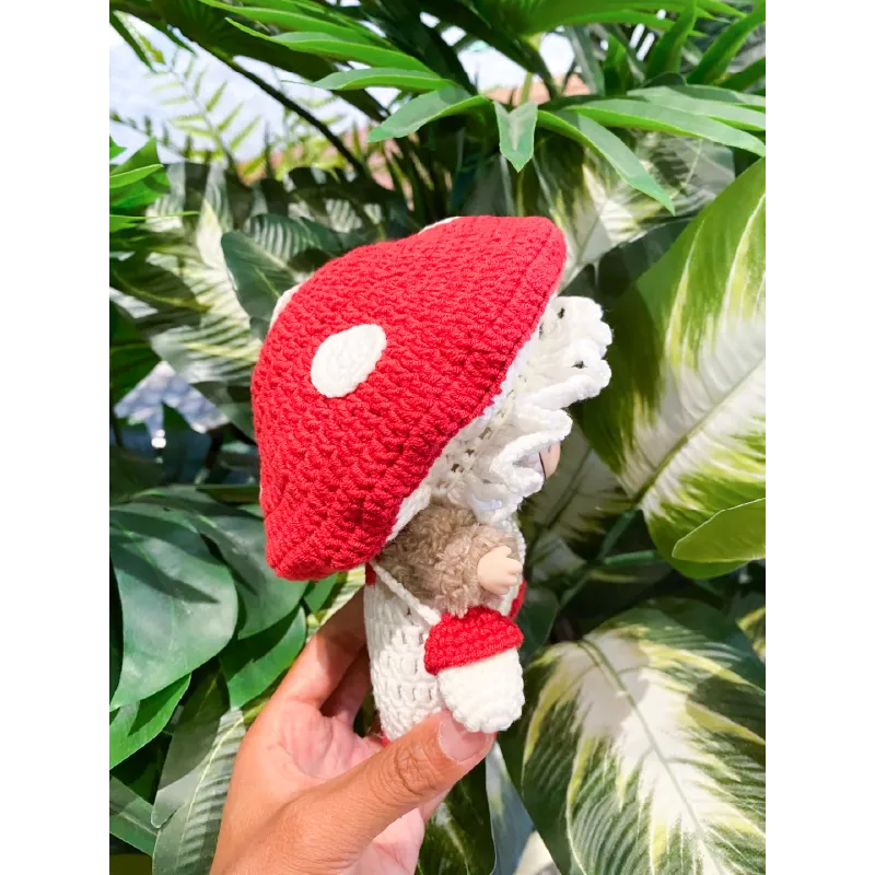 Hand holding a colorful Crochet Mushroom Doll with red cap and white spots surrounded by green leaves Crochet Mushroom Doll Pattern
