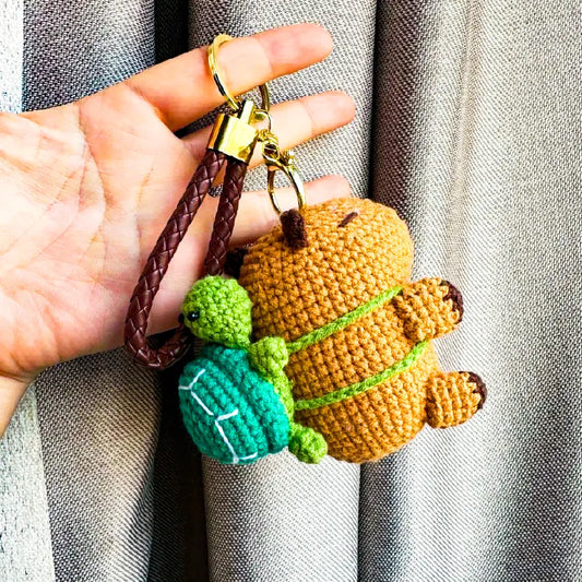 Hand holding crochet capybara keychain pattern with green turtle charm and braided leather loop