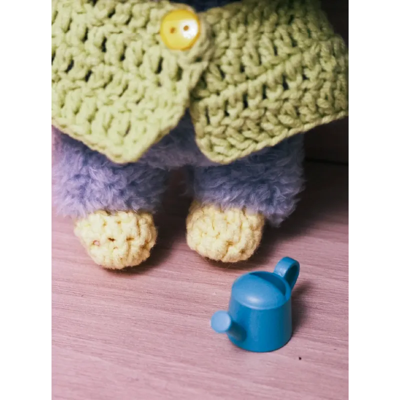 Close-up of frog knit doll outfit featuring crocheted green jacket and yellow booties with small blue watering can