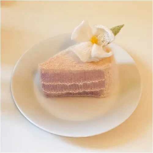 Knitted dessert pattern of a pink layered cake slice with a white flower on top on a white plate