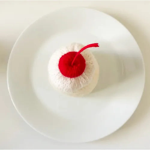 Knitted pudding pattern with white body and red cherry on top displayed on a white plate