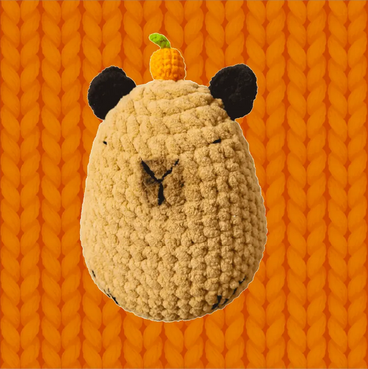 Round capybara crochet soft toy pattern featuring a beige plush with black ears and an orange decoration on top
