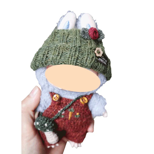 Handmade plush toy with a green knitted hat and red outfit, held in a hand against a white background.