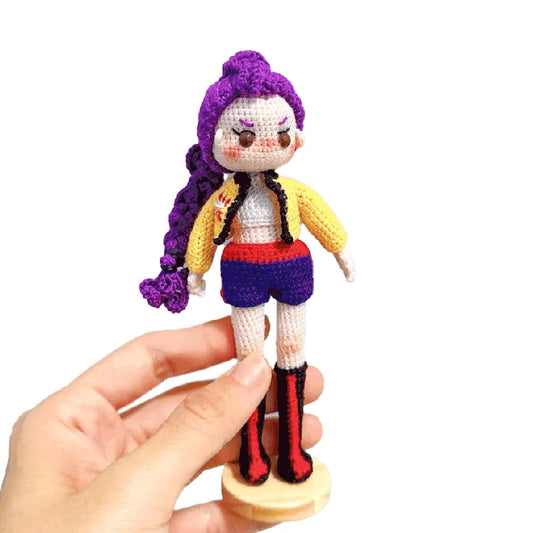 Hand holding a small crochet doll with purple hair and colorful outfit on a white background