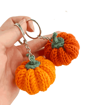 Hand holding two orange knitted pumpkin keychains with green stems on a white background